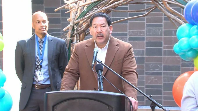 Mayor Bruce Harrell stands at podium in brown suit, speaking into microphone. Person in dark suit stands behind, multicolored balloons on both sides of podium