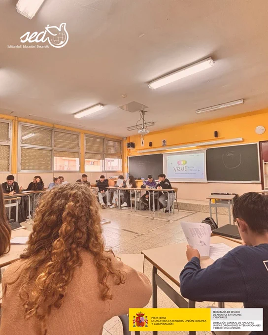 Un grupo de estudiantes de 4º de ESO de una escuela de Catalunya están sentados en círculo dentro de un aula escolar, participando en una actividad educativa. En la pizarra se proyecta el logo del proyecto "Veus per la infància" (Voces por la infancia). La iniciativa es llevada adelante por la ONGD SED (Solidaridad, Educación y Desarrollo), con el apoyo del Ministerio de Asuntos Exteriores, Unión Europea y Cooperación de España. 