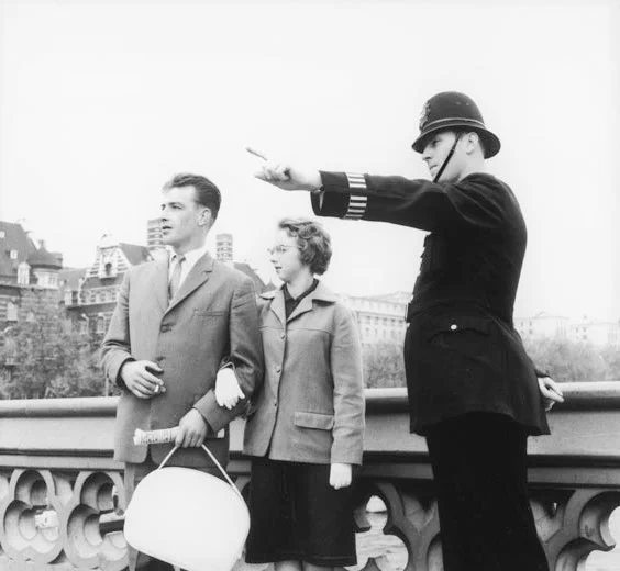 Black and white photograph of a male police officer pointing in the distance, with a couple standing next to him. The officer is wearing a striped armband on his arm.