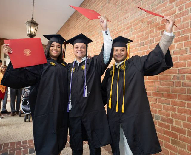Three graduates in black robes and honor cords celebrate by holding up their diplomas outside a brick building.