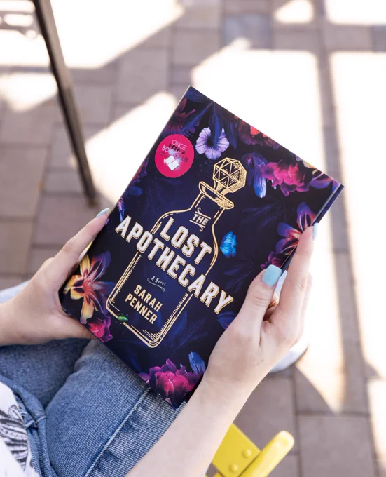 white hands hold a hardcover edition of The Lost Apothecary in their lap