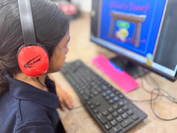 (Photo) A Miami Lighthouse Academy student wears headphones, and is learning computer skills in technology class by playing an educational game on the computer.
