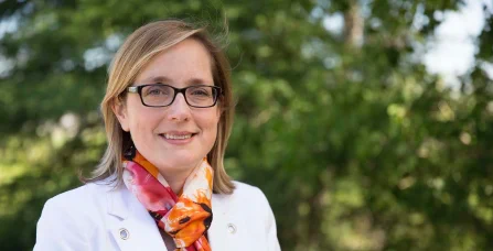 A photo of Dr. Janet DeMille, Medical Officer of Health and CEO of Thunder Bay District Health Unit. She is wearing a white blazer and colorful orange scarf and standing in front of some trees.