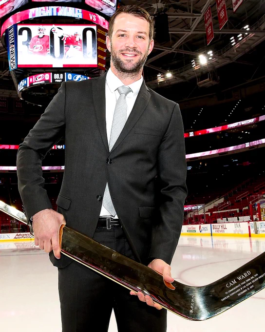 Cam Ward holds a commemorative goalie stick on ice in a black suit. 