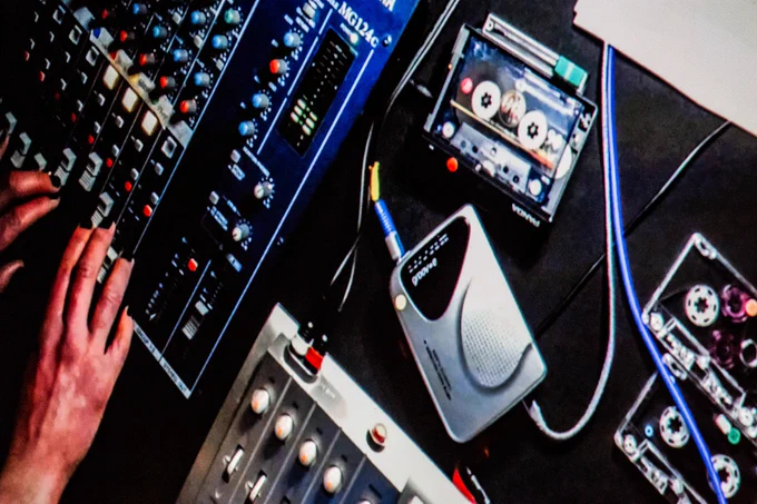 Photo of a webcam feed projected onto a large screen featuring slightly distorted footage of hands operating a mixer, plus several cassette tapes, a walkman and part of a 4-track tape machine visible.