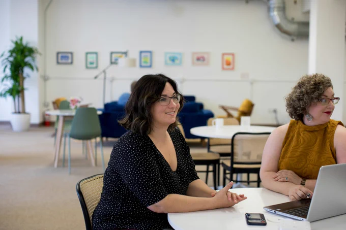 Two people who appear to be white women are sat at a round table in a bright, modern office space, smiling and looking over towards someone out of shot.