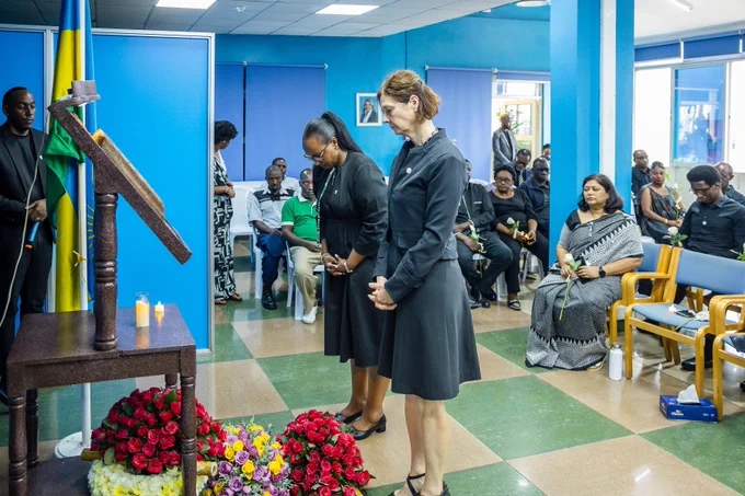 Two women, in black attire, bow in respect at a memorial stand bearing wreaths.
