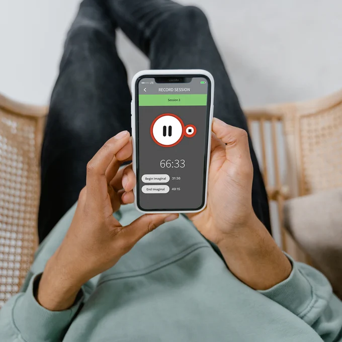 A person wearing a gray-blue hoodie and black jeans sits in a chair, holding a smartphone displaying the PE Coach mobile app interface.