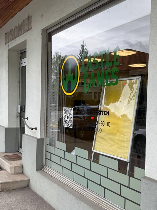 Picture of the entrance to the CastleGames store for trading card and video games in Oberburg, Switzerland. Shown in the foreground is the main display window with the stores opening hours on Tuesdays to Thursdays from 18:00 to 20:00 and on Fridays from 18:00 to 22:00.