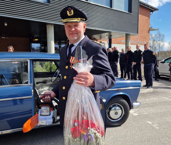 Pelastusalan juhlapukuun pukeutunut mies iloisena kukat kädessä. Takana vanha auto.