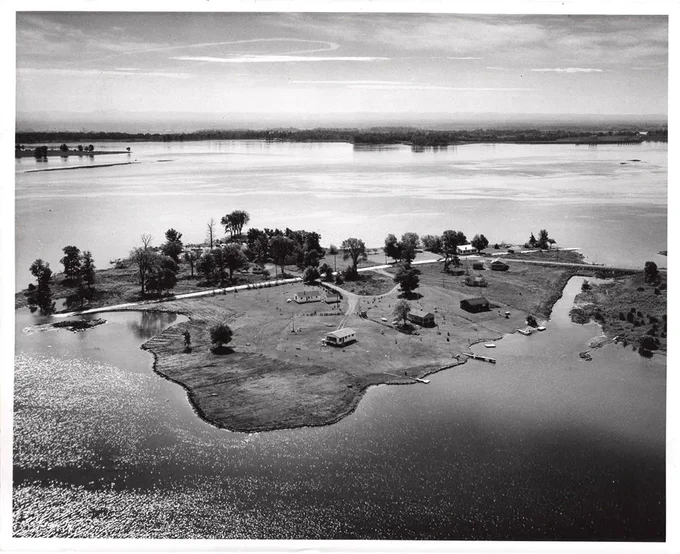 A black-and-white aerial photo shows a small island surrounded by a large body of water. The island has scattered trees, a few buildings, and open grassy areas, with a narrow waterway cutting through one side. The horizon reveals a distant shoreline under a partly cloudy sky.