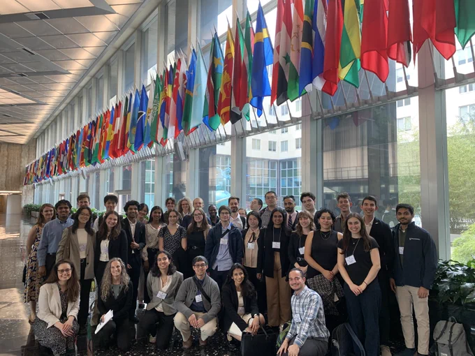 Group of young professionals and students posing for a photo inside the U.S. Department of State, with an array of international flags hanging above them along a large windowed hallway. 