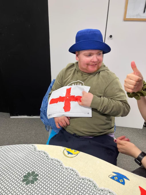 A young man in a khaki green hoodie and blue bucket hat holding up a homemade St George's cross. 