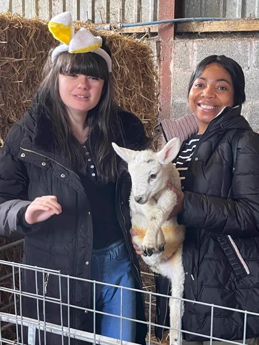 Two young women both wearing black coats hold a white lamb. Behind them are stacks of straw bales. The woman on the left is wearing bunny ears on her head. 