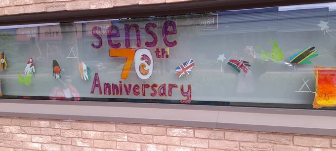 A window with 'Sense 70th Anniversary' written in orange and purple chalk pen. Around the window are white shooting stars and hand prints decorated with different colours. 