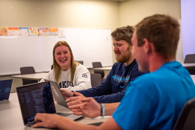 Three students, one wearing a Dakota State sweatshirt and another in a "Trojans" t-shirt, are engaged in a lively discussion around a laptop in a classroom.