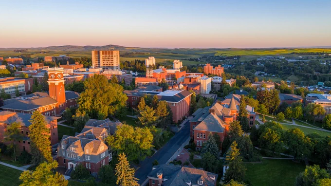 Color aerial photo of the W S U Pullman campus at sunset. Various red brick buildings are surrounded by green fields and green trees. 