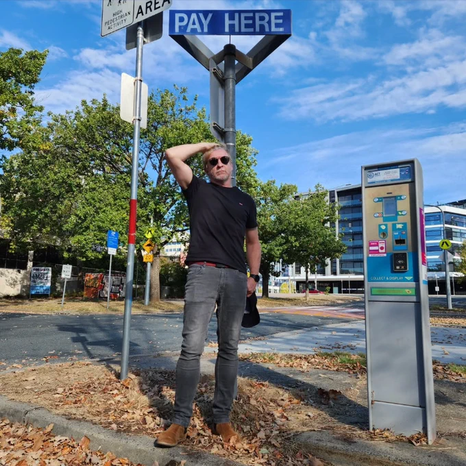 Rhys darby stands at a parking meter with a sign that says Pay here. He has his hand on his head, and wearing a black shirt and grey jeans. 
