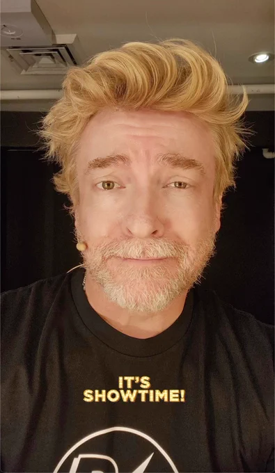Rhys Darby looks at the Camera with Blonde, gravity defying hair, and a cinnamon sugar colored beard. The man is sunshine personified. There is yellow and white text that says "It's showtime!"
