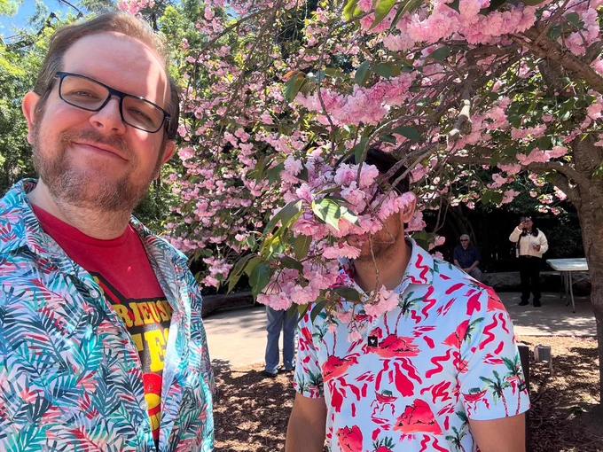 Dominic Burgess stands in front of a cherry blossom tree with his husband hiding behind said blossoms