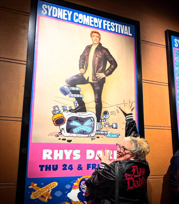 Gypsy Taylor Stands in front of a huge Sydney Comedy Festival poster with Rhys Darby standing on a robot. She is wearing her Our Flag Means Daddy Jacket, smiling. 
