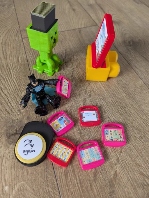 Photo of two plastic toys, a green Minecraft creeper and tiny batman, surrounded by 3D printed miniature AAC devices.