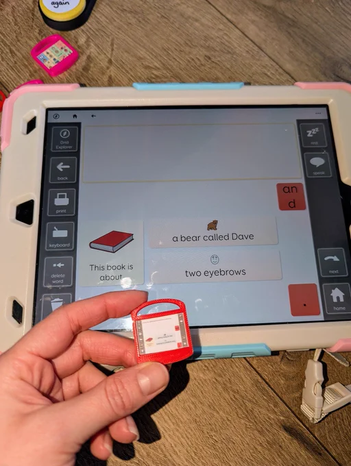 Photo of an iPad displaying a CALL Scotland Bookbug Grid writing grid. A hand in the bottom left corner is holding a 3D printed miniature iPad with a printed screenshot of the same writing grid.