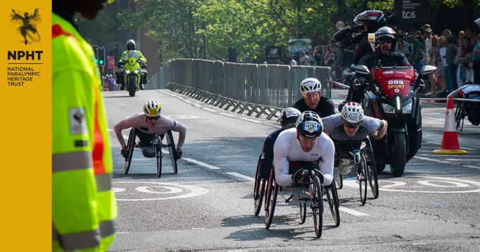 A group of 4 wheelchair athletes wearing helmets and battling it out for position on the road, with a motorbike and camera following them. Sean Frame leads, his eyes focussed on the road ahead. Behind him is Evan Correll, then Nathan Maguire with his race goggles on and grimacing, and Hiroki Nishida wearing see-through racing glasses and his mouth open to take a breath. A fifth athlete is pictured further behind them, followed by a police motorbike. Left of frame is a marshal wearing a high vis yellow jacket.