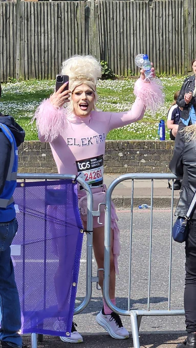 Elektra Fence running in drag, wearing a pink bodysuit with fluffy sleeves - smiling and cheering running past the Scope cheerpoint