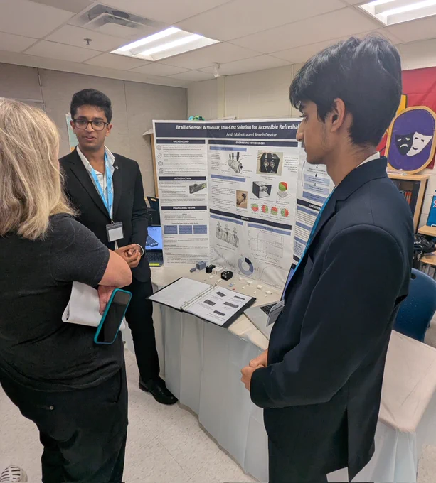 Two male students in suit blazers stand in front of a presentation trifold. They are speaking to an adult female.