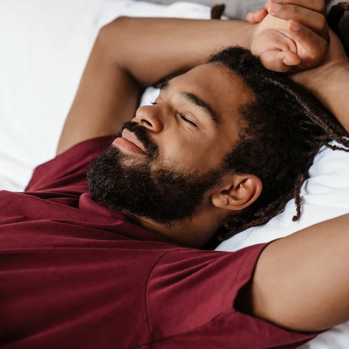 A man with a beard wearing a red t-shirt, sleeps in bed, stretching his arms above his head.