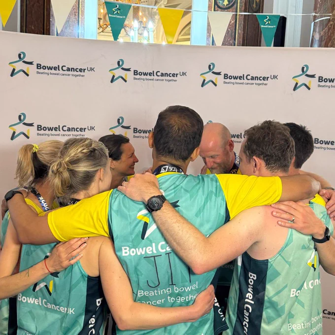 A photo of a team of London Marathon runners in a huddle with their arms around each other. They're wearing Bowel Cancer UK branded t-shirts. The photo features a Bowel Cancer UK branded wall and bunting.