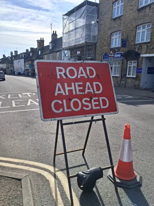 Red sign with white text reading "Road Ahead Closed" with a traffic cone beside it.