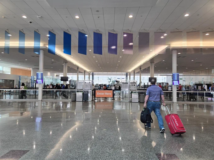 Photo shows passenger with a red rolling suitcase walk toward the Tulsa International Airport's TSA Security checkpoint for screening. Above the entrance of the checkpoint, there is a colorful hanging art display that shows over a dozen panels in a variety of colors. 
