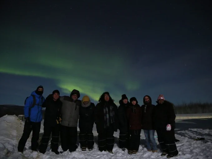 A row of people bundled well against the cold, stand on snow beneath the green light of the aurora