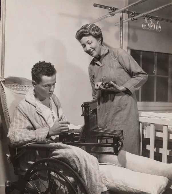 Injured soldier in a wheelchair, being taught to weave by a woman