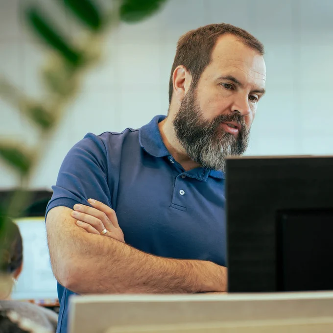 Person in a blue polo shirt standing with arms crossed, looking at a laptop screen with a thoughtful expression in an office setting.