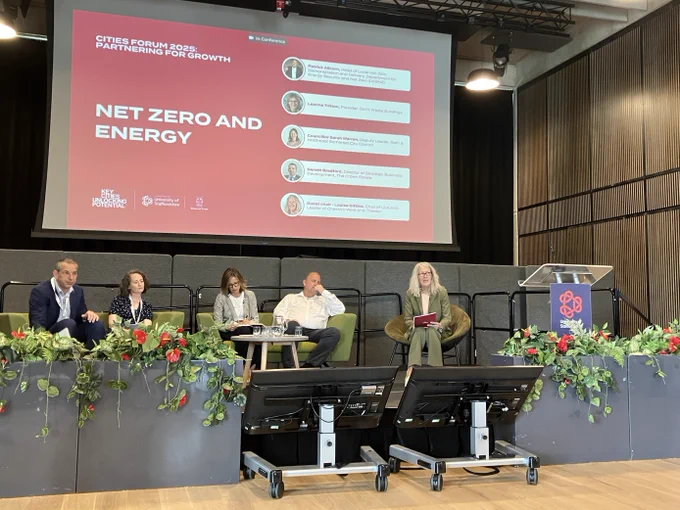 A panel of five speakers seated on stage at the 'Cities Forum 2025: Partnering for Growth' conference, under a large red presentation screen titled 'Net Zero and Energy.' The event is hosted by the University of Staffordshire, with a podium decorated with flowers and conference branding in front of the stage