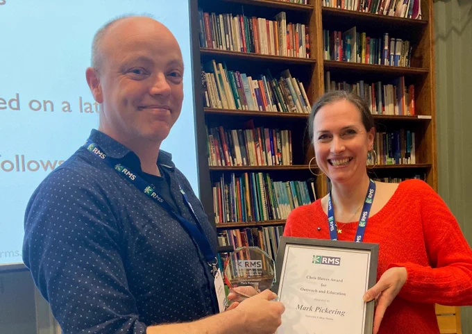 Mark Pickering (left, smiling, wearing dark shirt) receiving the RMS Chris Hawes Award and certificate for Outreach and Education from Kerry Thompson (smiling, wearing red top). A book case and projector screen are in the background.