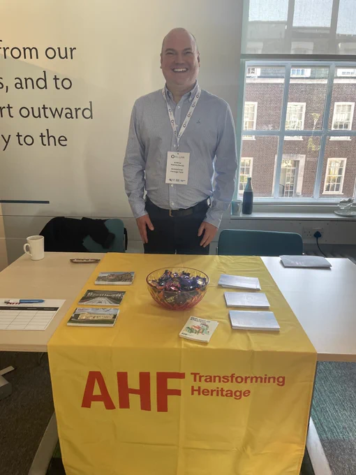 AHF Investment Manager, Andrew Hitches-Davies, at the Good Finance Live event in London. Andrew is standing behind a table, which is covered in a yellow runner with the red AHF logo on it at the front. There are some postcards, leaflets, and a bowl of sweets laid out on the table.