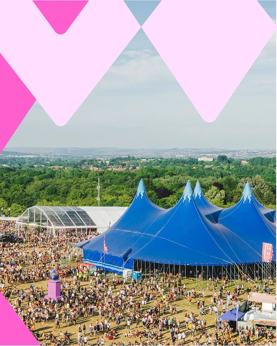 Outdoor music festival with large blue tent, crowds, and greenery in the background.