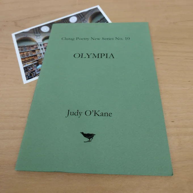 A close shot of a slim green poetry  book published by Clutag Poetry New Series No. 10 titled Olympia by Judy O'Kane sits on top of a partially obscured postcard showing a grand library. These sit on a wooden desk.