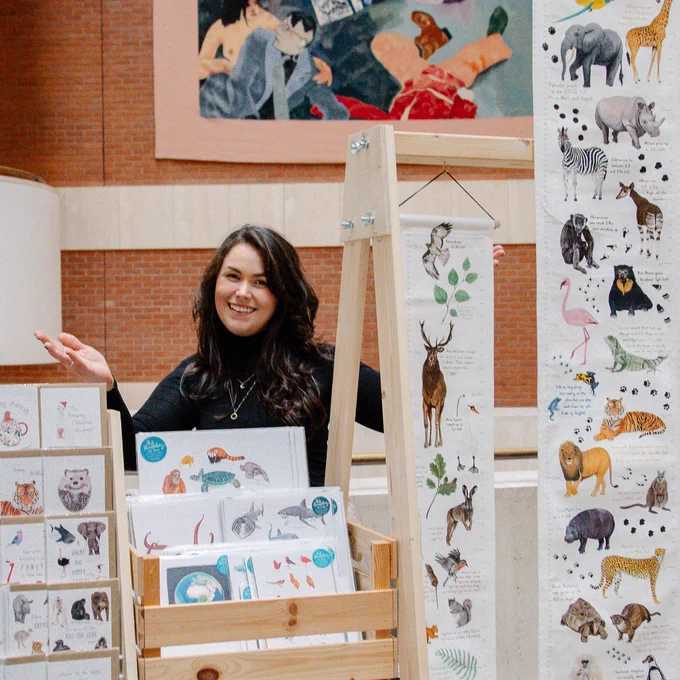 A photo of a market trader smiling with their handmade illustrations at an indoor market.