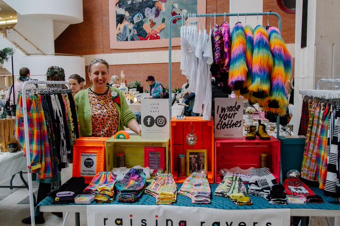 A photo of a market seller standing behind a table of their handmade colourful clothing in an indoor craft market.