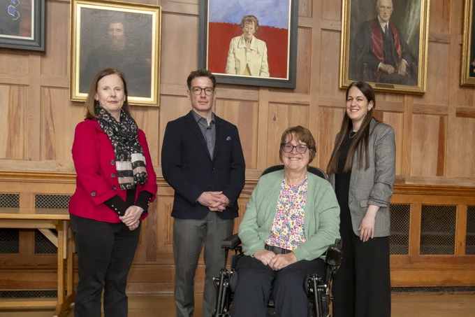 Image shows Professor Denise Fitzgerald, Principal Investigator and researcher from the Wellcome-Wolfson Institute for Experimental Medicine at Queen’s University Belfast; Stewart Finn, Country Director at MS Society Northern Ireland; Caroline Millar who is living with MS; and Dr Emma Gray, Director of Research at the MS Society. They are pictured beside a portrait of Professor Dame Ingrid Allen in the Great Hall at Queen's.