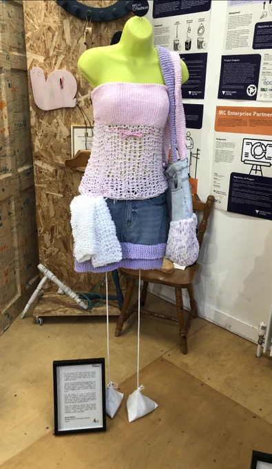 A mannequin dressed in a sustainable fashion outfit created for the ReLove Fashion competition. The outfit features a handmade lavender and white crochet-style strapless top, paired with a denim mini skirt that has a purple knitted trim at the hem. Accessories include a chunky knitted shoulder bag and a white textured scarf or towel draped over the left hip. The mannequin stands on weighted strings, and a framed description of the project is displayed on the floor in front. 