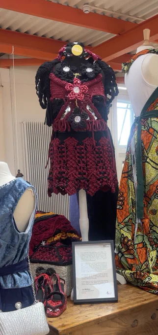 A mannequin displays a bold, handcrafted outfit for the ReLove Fashion competition, featuring intricate crochet work in deep red and black. The outfit includes a fitted bodice with decorative mirror elements and floral detailing, layered over a black fabric base. The skirt has a scalloped hem with thick woven patterns. A framed project description is placed on the floor in front of the mannequin. Surrounding the mannequin are other colourful garments, including  matching accessories like a pair of red handcrafted shoes.