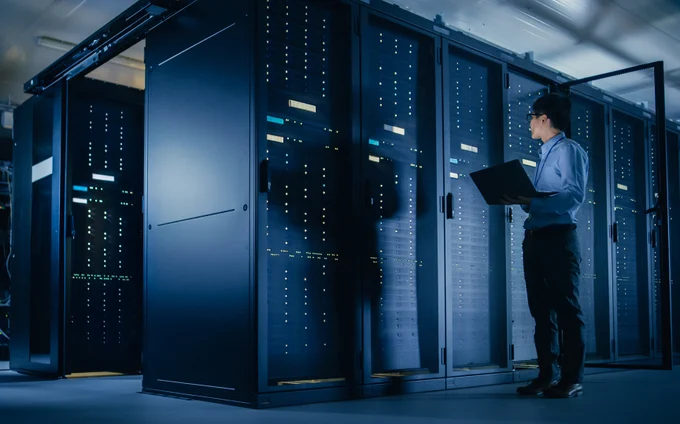 In a data center, a male IT Technician stands and looks at a laptop next to a server rack. Credit: Getty Images