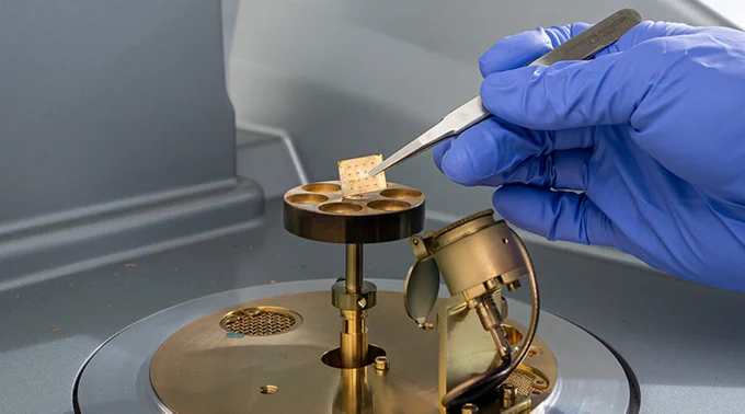A blue, gloved hand holds a small, square, gold film over an ion sputtering machine.