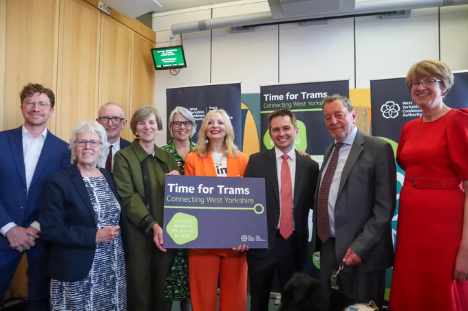 Group of nine people, stood, smiling. In the middle, they are holding a sign that says Time for Trams, Connecting West Yorkshire.